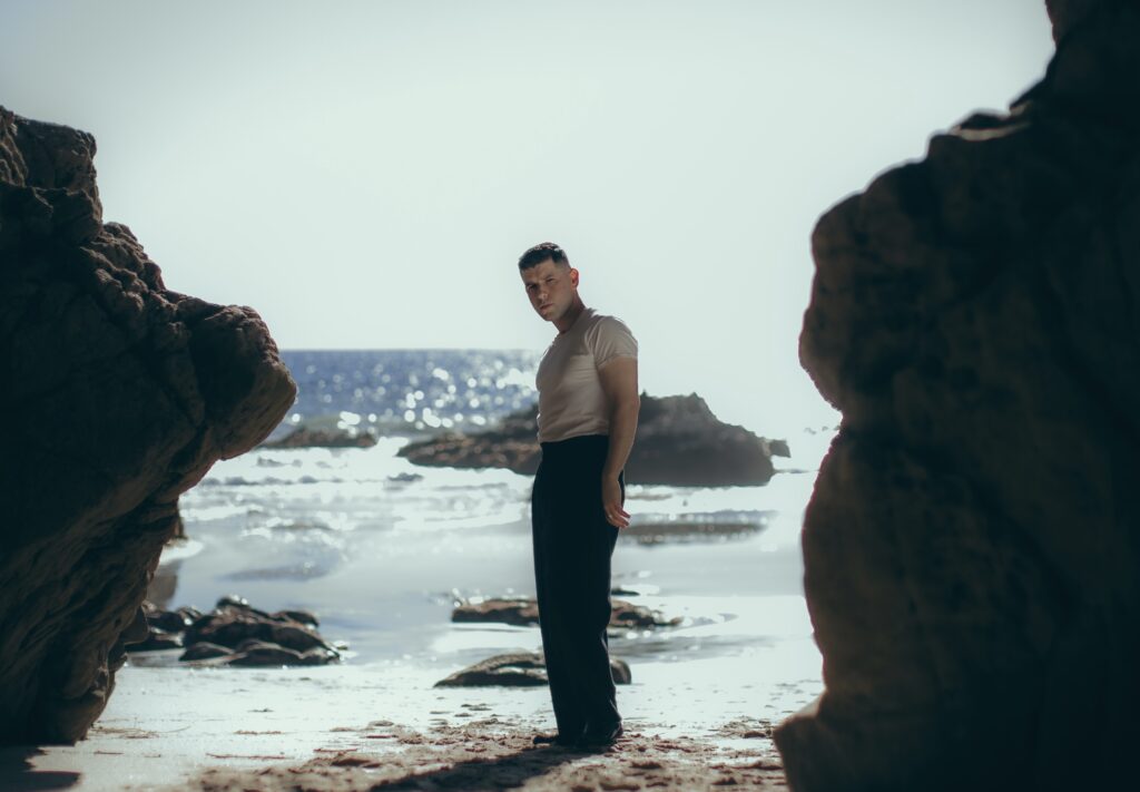 Singer Aaron Frazer stands in front of a tranquil view of the ocean with vibrant blue water and a cloudless sky.