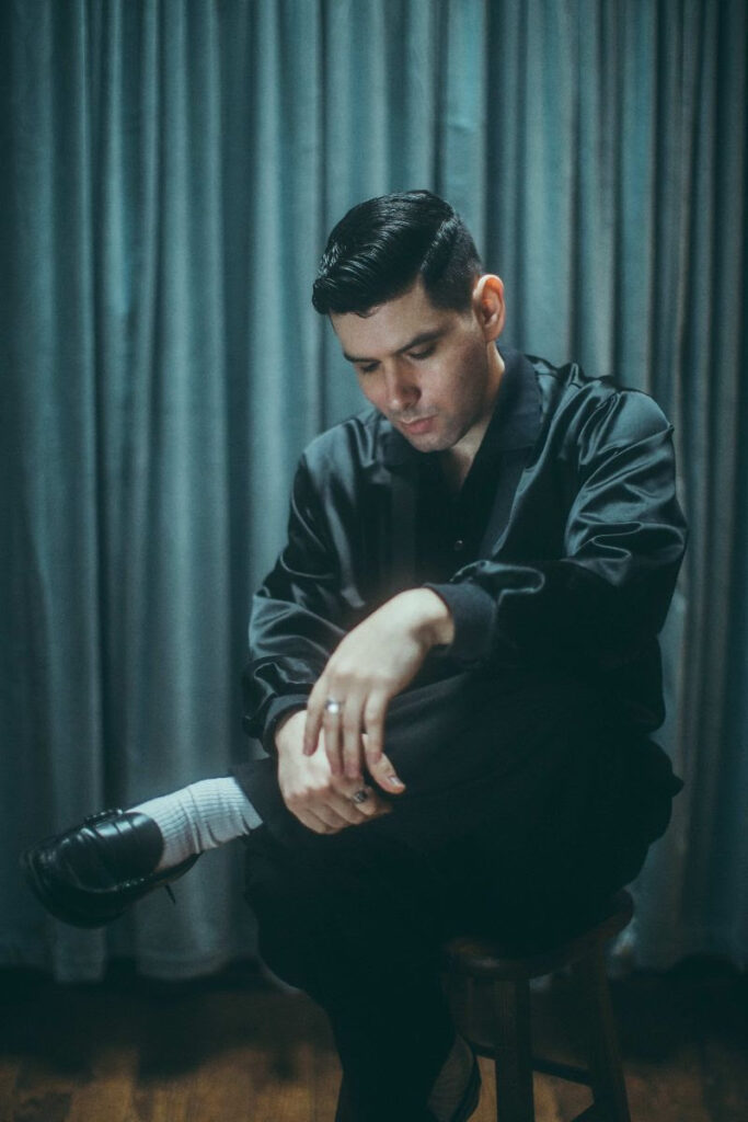 Image of Aaron Frazer in a black jacket sitting on a stool.