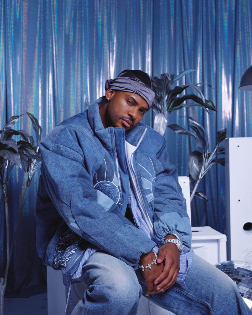 Trevor Jackson sits casually in a stylish denim outfit, accessorized with a bandana and silver jewelry, against a backdrop of shimmering metallic curtains, exuding a relaxed, fashionable vibe.