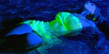 The cover art for Chloe Bailey's album Trouble in Paradise. She is floating in water, illuminated by blue and green lights, wearing a glow-in-the-dark costume resembling a mermaid tail and bikini top. The reflective details on the costume sparkle subtly under the light.