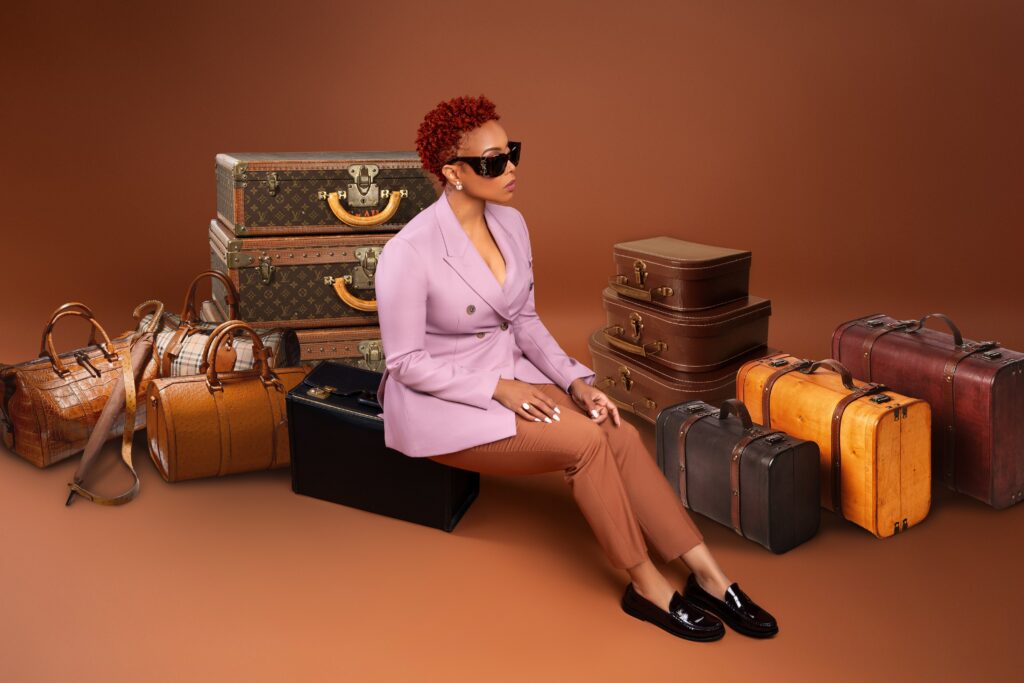 R&B singer Chrisette Michele with short red hair, wearing sunglasses, a lavender blazer, and tan trousers sits on a black suitcase among a collection of various vintage suitcases in rich browns and tan colors. The setting has a warm, monochromatic brown background.