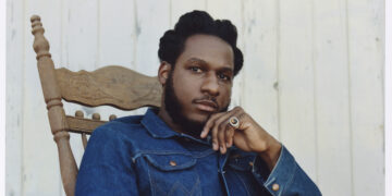 A photo of singer Leon Bridges, wearing a blue denim jacket, sitting in a rocking chair