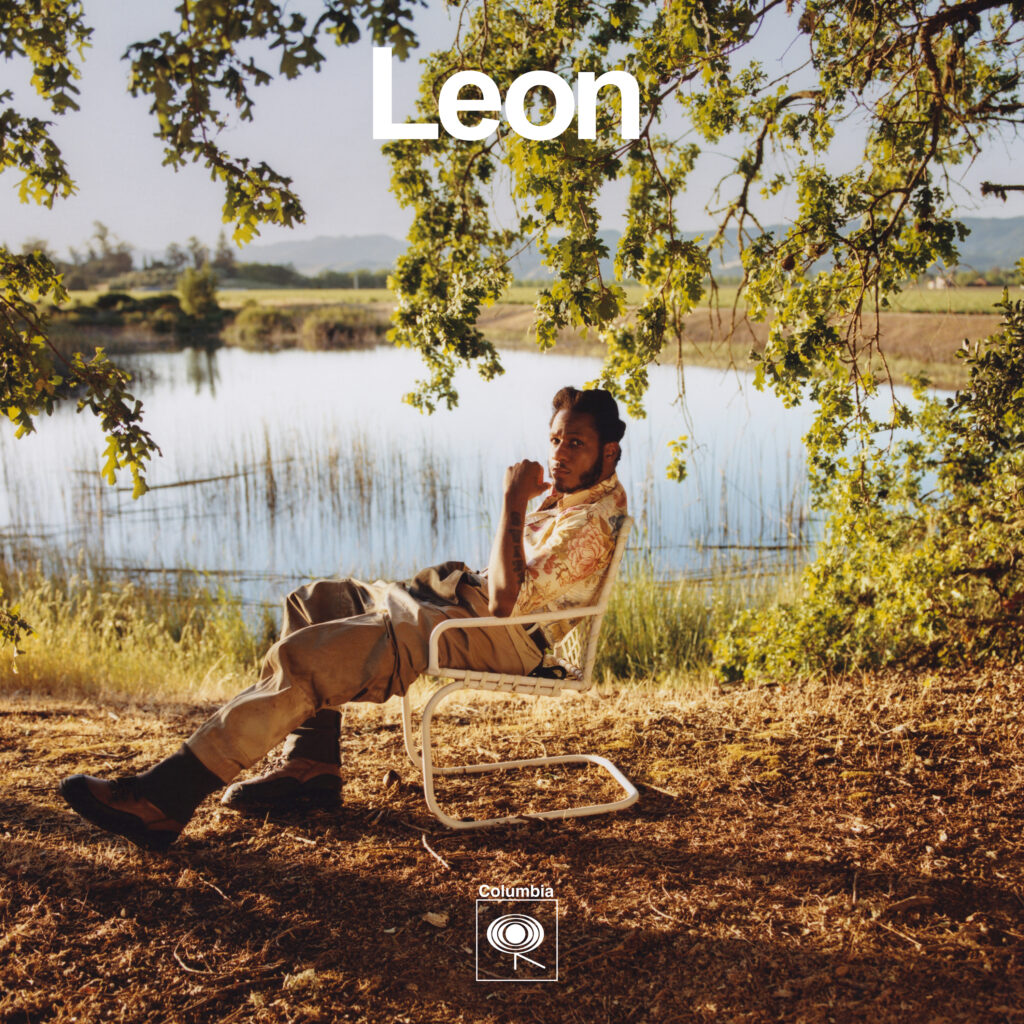 The image shows singer Leon Bridges in a relaxed pose on a folding chair next to a calm lake, surrounded by grass fields and trees under a clear sky. He is wearing a casual, patterned short-sleeved shirt and light trousers, one hand thoughtfully touching his chin. In the upper left corner is the text "Leon" appears in large letters. In the lower right corner is the Columbia brand logo.