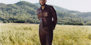 A photo of singer Leon Bridges standing in a sunlit field with tall grass, wearing a stylish leather jacket and gazing off into the distance. A forest and a small hill are visible in the background under a clear sky.