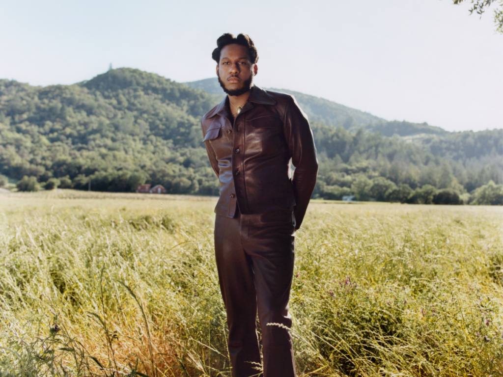 A photo of singer Leon Bridges standing in a sunlit field with tall grass, wearing a stylish leather jacket and gazing off into the distance. A forest and a small hill are visible in the background under a clear sky.